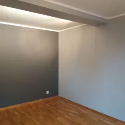 Pomalowane na szaro wnętrze pokoju z drewnianą podłogą; jedna ściana ciemnoszara, druga jasnoszara, z widocznymi gniazdkami elektrycznymi.