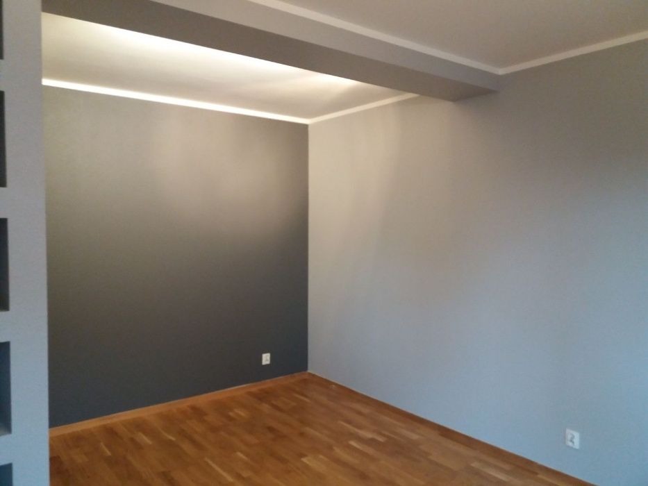 Pomalowane na szaro wnętrze pokoju z drewnianą podłogą; jedna ściana ciemnoszara, druga jasnoszara, z widocznymi gniazdkami elektrycznymi.