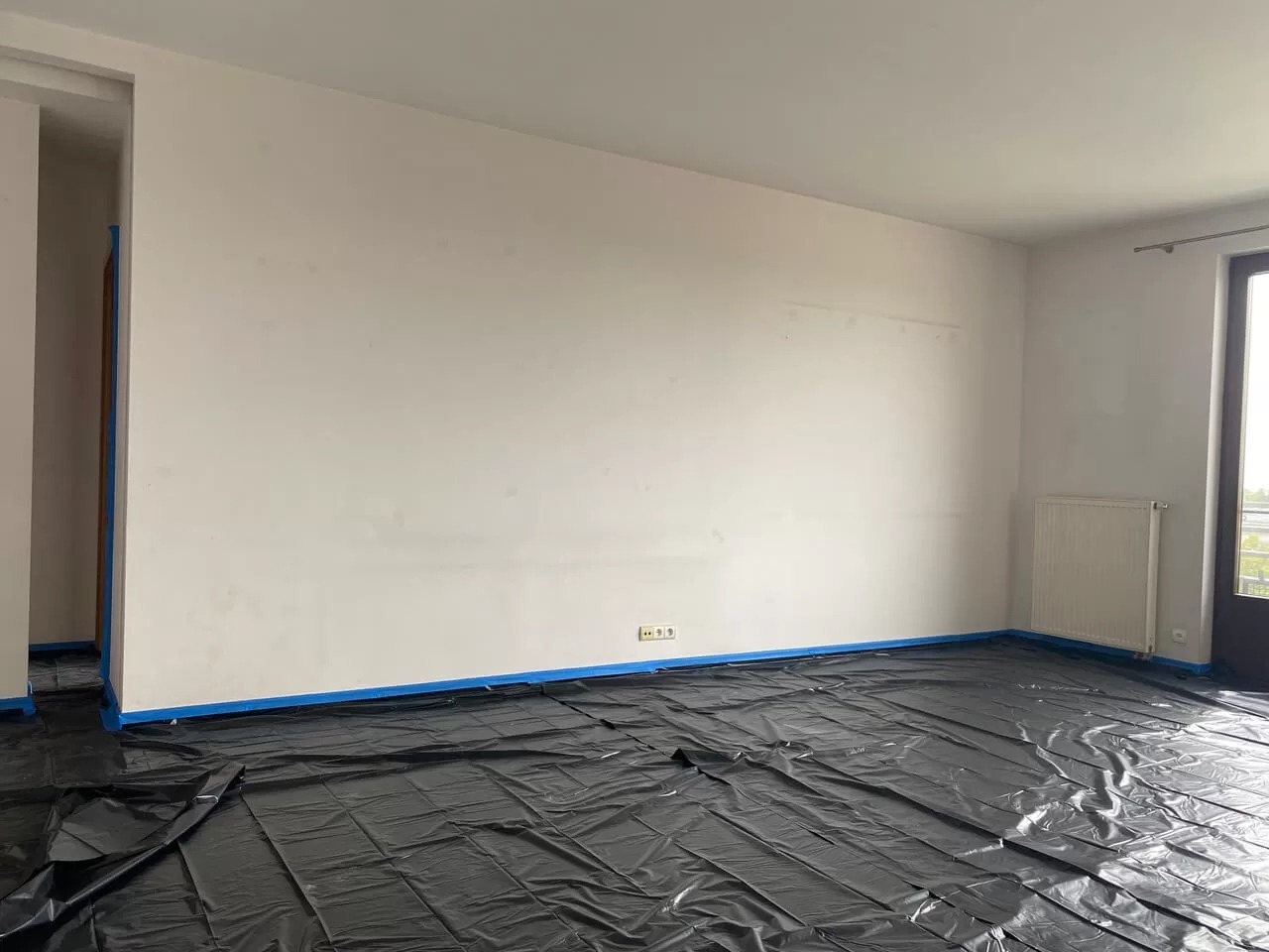 Przygotowanie pokoju do malowania: podłoga zabezpieczona czarną folią, ściany i framugi oklejone niebieską taśmą malarską, widoczne gniazdka elektryczne i grzejnik.