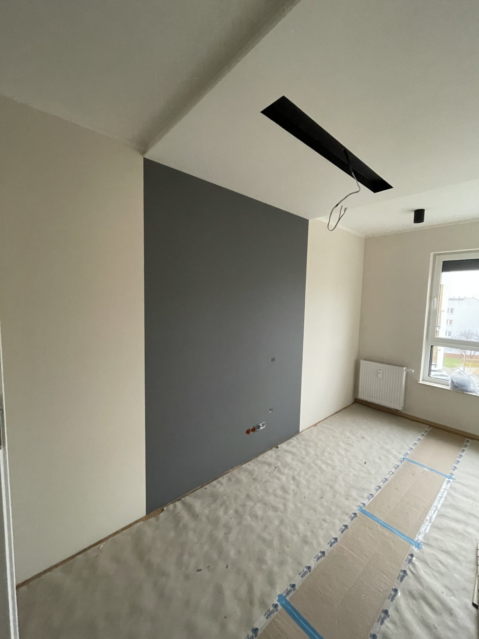 Świeżo pomalowane wnętrze w Kaliszu z szarą ścianą akcentującą, zabezpieczona podłoga, widoczne gniazdka elektryczne i przygotowane oświetlenie sufitowe.