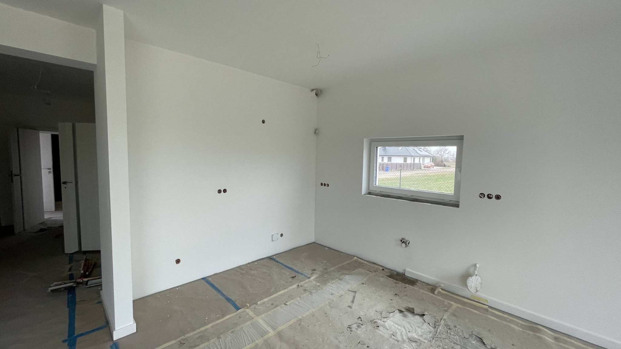 Wnętrze pokoju w trakcie wykańczania: świeżo pomalowane ściany na biało z widocznymi otworami na gniazdka elektryczne, okno z widokiem na zewnątrz, podłoga zabezpieczona folią malarską, widoczny...