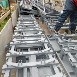 Szare, spawane konstrukcje metalowe z uchwytami, ułożone rzędami; robotnik w czerwonej koszulce i sandałach stoi na jednej z nich.