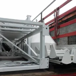 Konstrukcja stalowa pomalowana na biało, przewożona na platformie transportowej z czerwonymi elementami zabezpieczającymi.