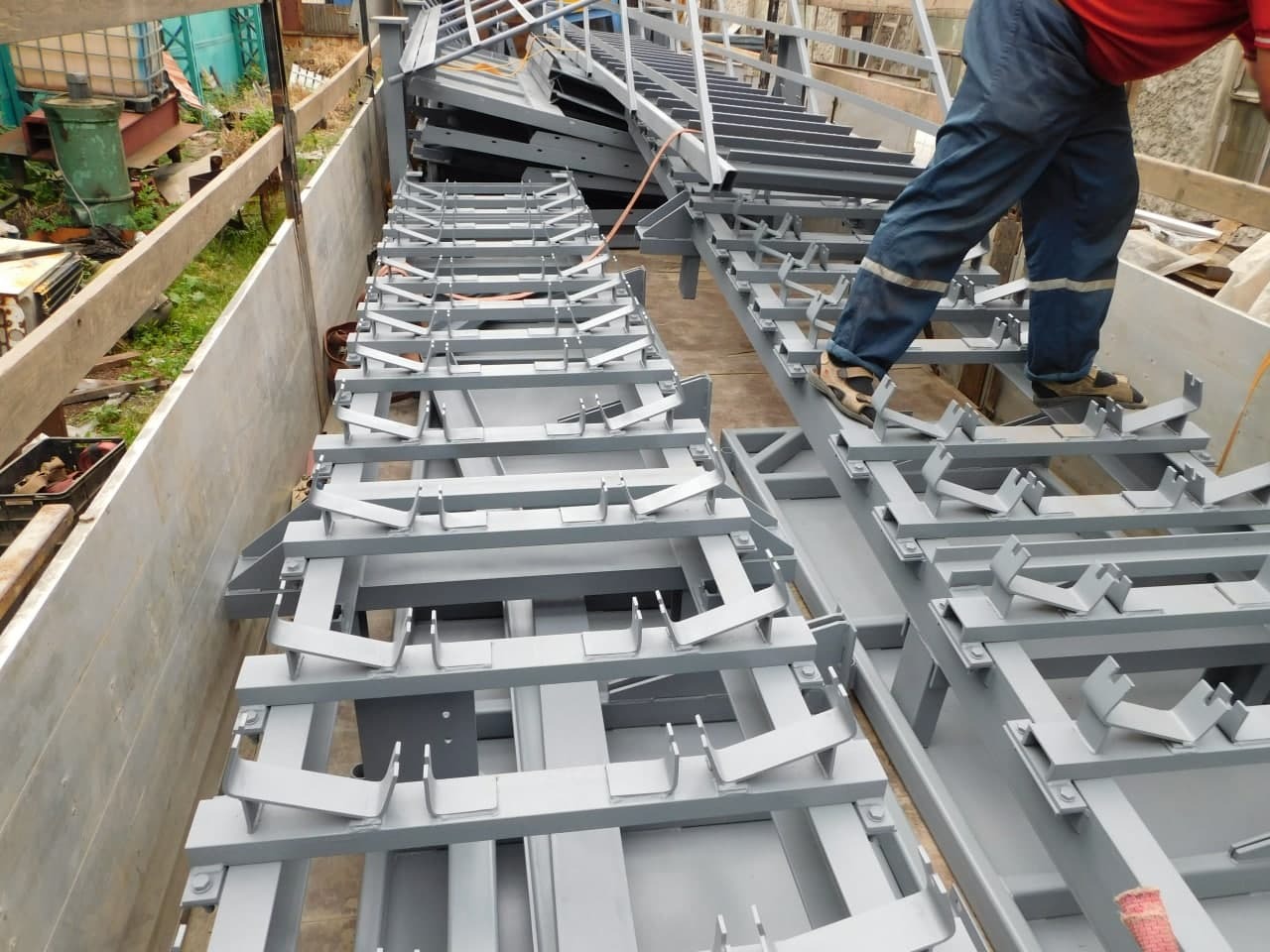 Szare, spawane konstrukcje metalowe z uchwytami, ułożone rzędami; robotnik w czerwonej koszulce i sandałach stoi na jednej z nich.