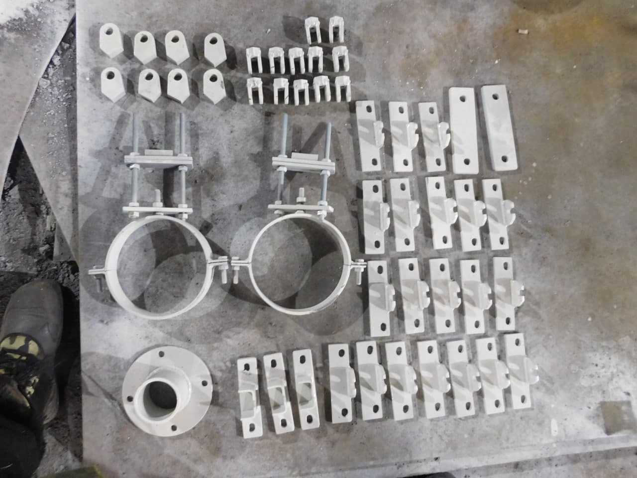 Białe elementy konstrukcyjne, obejmy rurowe z mocowaniami, uchwyty i złączki, rozłożone na szarej, betonowej powierzchni roboczej.