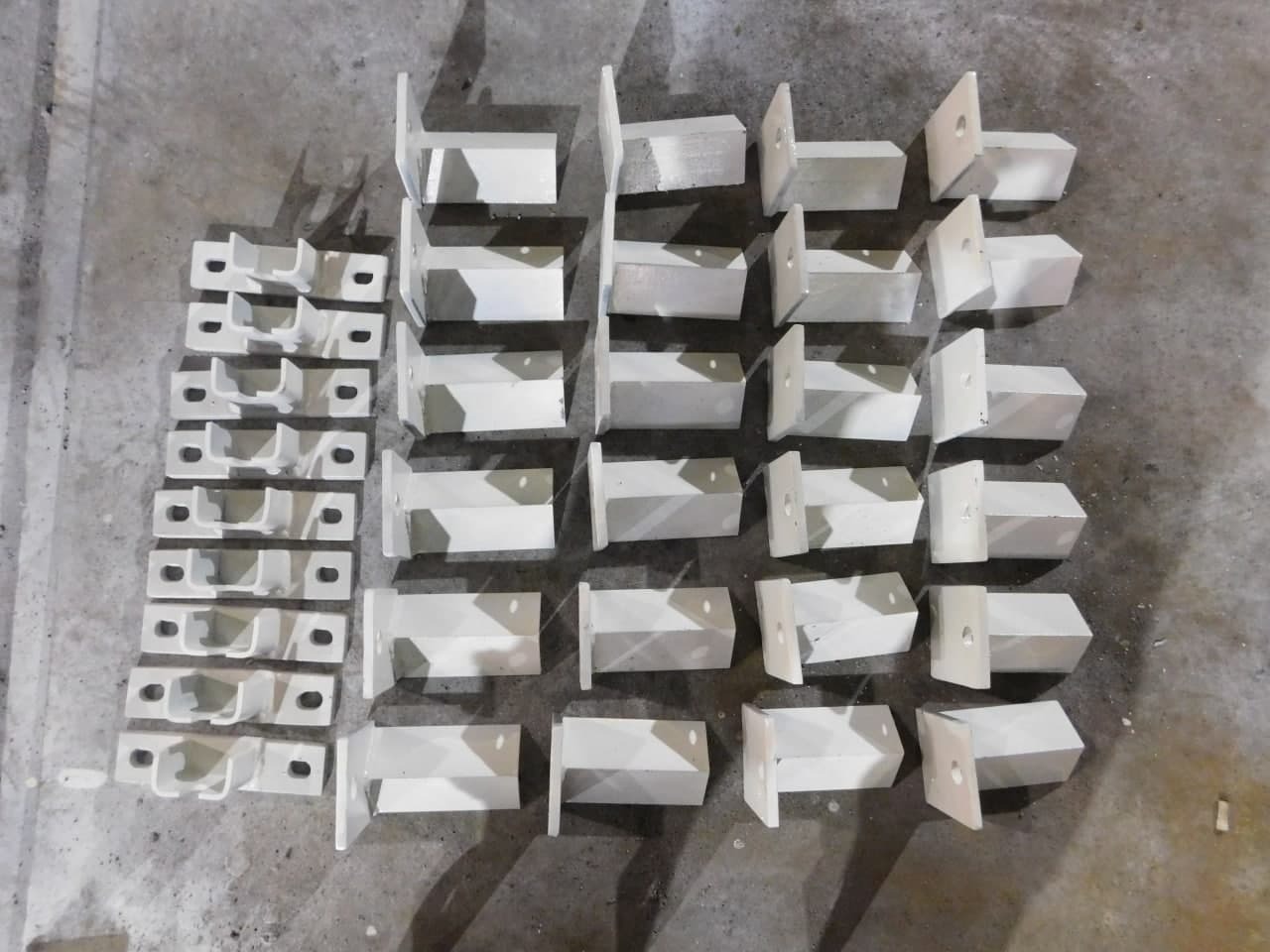 Partia pomalowanych na biało metalowych elementów konstrukcyjnych o różnych kształtach, ułożonych na szarej, betonowej posadzce.