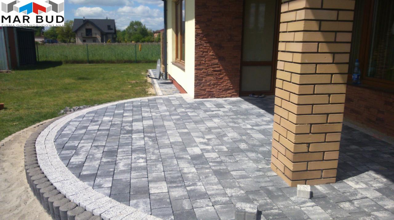 Wykończony taras z szarej kostki brukowej ułożonej w łuk przy domu z ceglaną kolumną, widok na trawnik i dom w tle.
