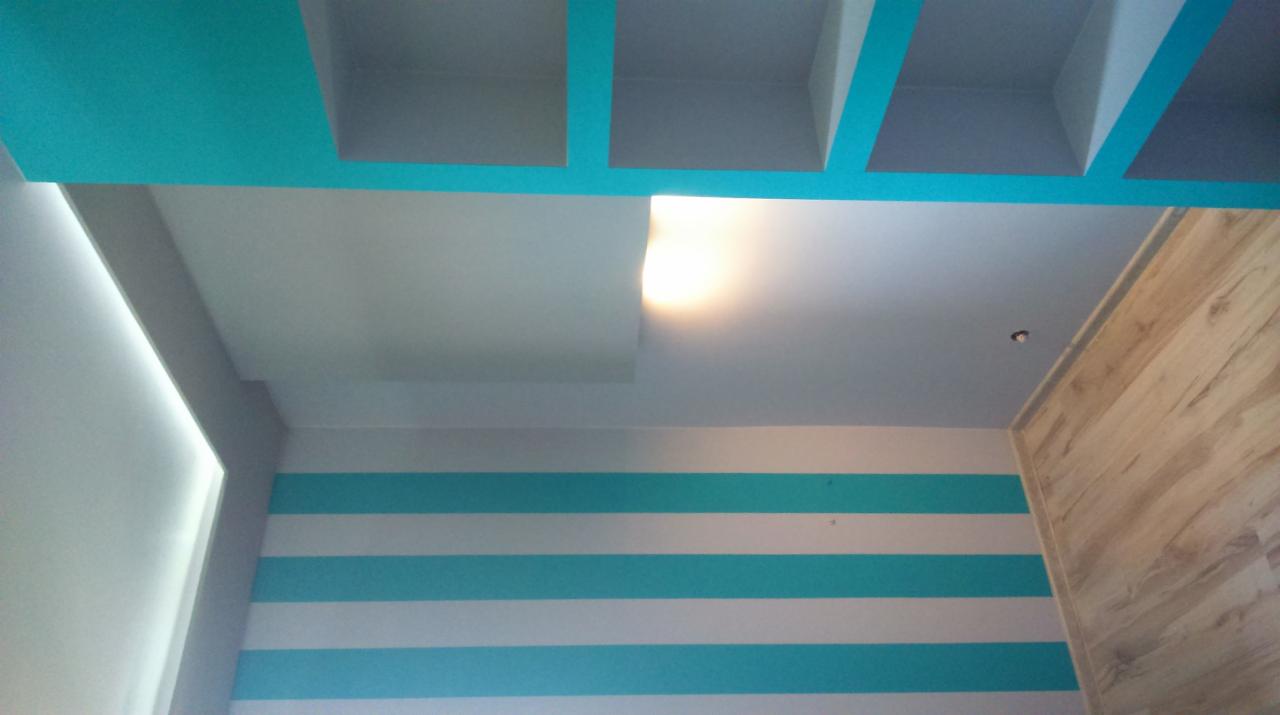 Wnętrze pokoju z dekoracyjnym sufitem podwieszanym z turkusowymi elementami, ścianą w poziome turkusowo-białe pasy i okładziną drewnopodobną na ścianie po prawej stronie.