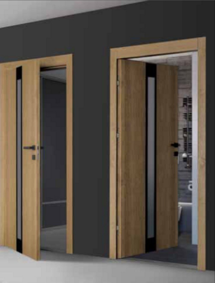 Dwa drewniane skrzydła drzwiowe z czarnymi wstawkami, jedno lekko otwarte ukazujące szarą ścianę, drugie otwarte na łazienkę z widoczną wanną i grzejnikiem.