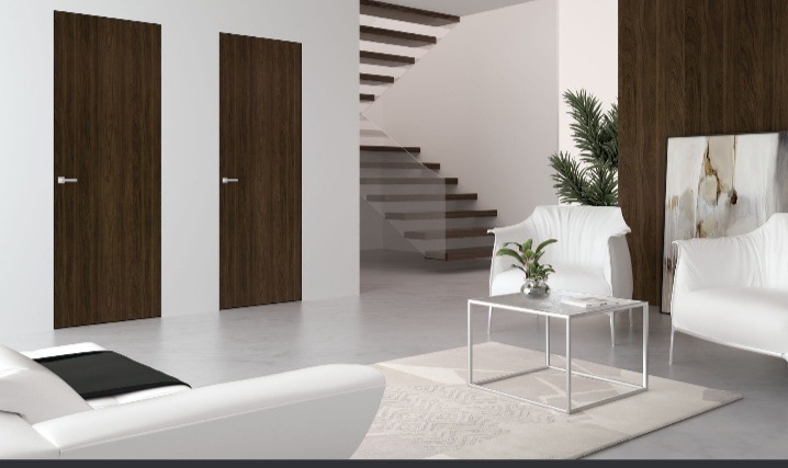 Dwie ciemnobrązowe, minimalistyczne drzwi wewnętrzne z srebrnymi klamkami, na tle jasnego salonu z białymi meblami i schodami.