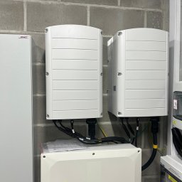 SolarGlek - Odnawialne Źródła Energii - Dwa białe inwertery SolarEdge zamontowane na szarej ścianie z betonowych bloczków, z widocznymi kablami i skrzynką przyłączeniową pod spodem. Na skrzynce ostrzeżenie o napięciu i logo SolarEdge.