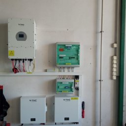 SolarGlek - Odnawialne Źródła Energii - Instalacja fotowoltaiczna wewnątrz budynku: białe falowniki V-TAC i skrzynki zabezpieczeń przymocowane do ściany z widocznymi przewodami i elementami instalacji elektrycznej.