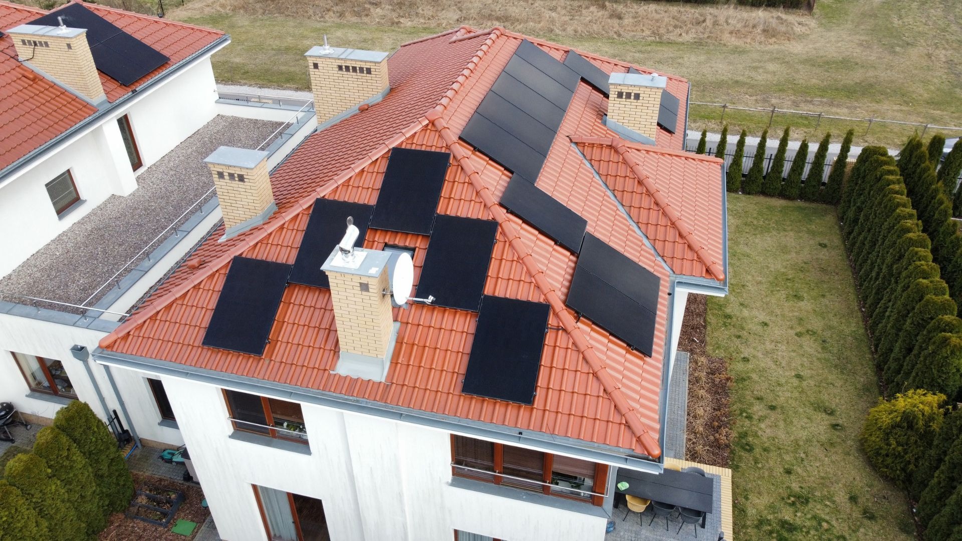 Widok z góry na dom z czerwoną dachówką, na której zainstalowano panele słoneczne; komin z anteną satelitarną.