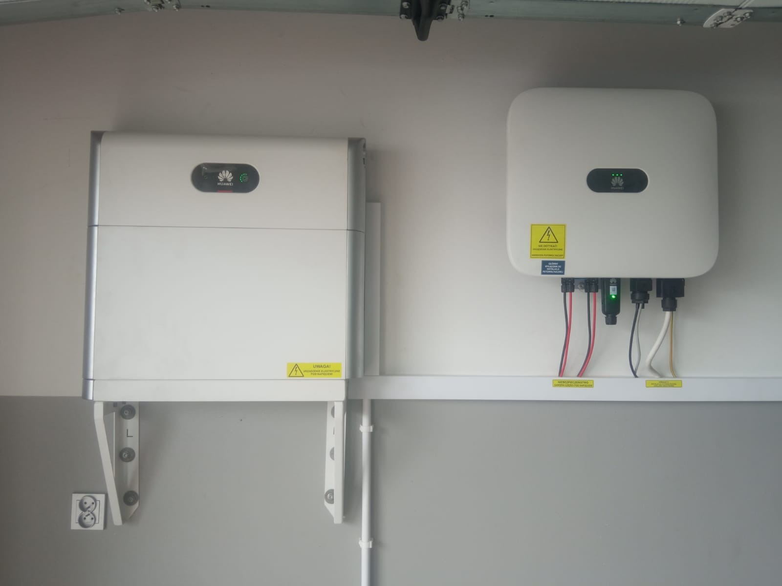 Biały magazyn energii Huawei po lewej i falownik Huawei po prawej zamontowane na ścianie garażu, widoczne kable i ostrzeżenia.