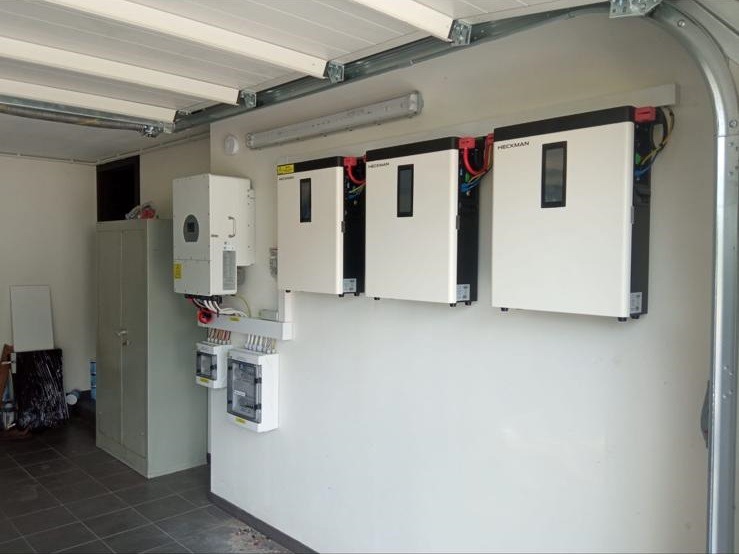 Instalacja magazynów energii Heckman w garażu, widoczne trzy białe urządzenia na ścianie, po lewej falownik i rozdzielnia elektryczna, metalowa szafa w tle.