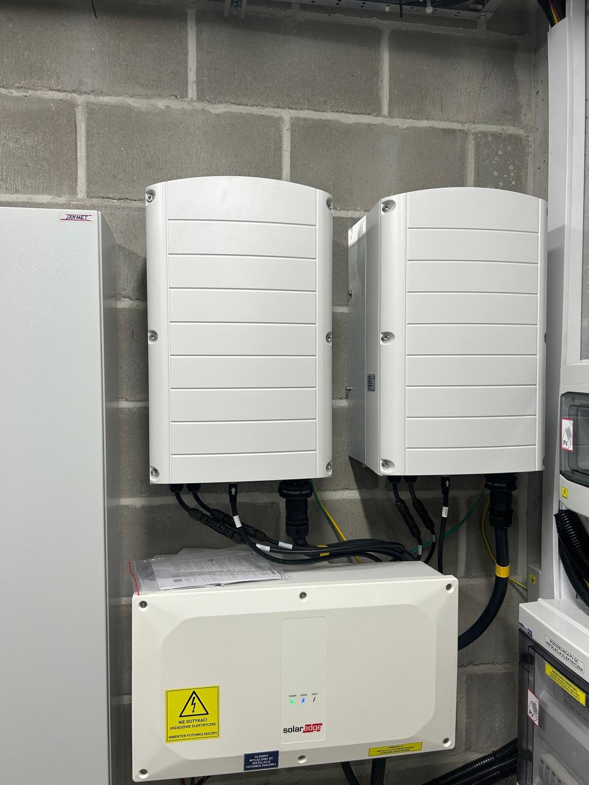 Dwa białe inwertery SolarEdge zamontowane na szarej ścianie z betonowych bloczków, z widocznymi kablami i skrzynką przyłączeniową pod spodem. Na skrzynce ostrzeżenie o napięciu i logo SolarEdge.