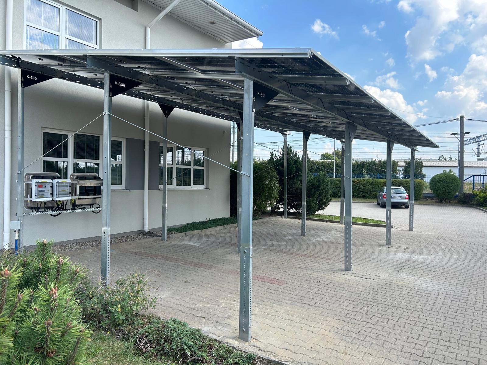 Zadaszenie parkingowe z panelami słonecznymi, szara elewacja budynku w tle, instalacja elektryczna na ścianie, kostka brukowa na parkingu.