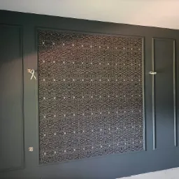 Ciemnozielona ściana z dekoracyjnymi panelami i wzorzystą tapetą w ramie, widoczne wystające kable elektryczne.