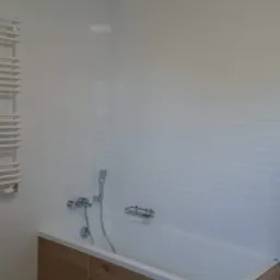 Wykończona łazienka z wanną obudowaną panelami imitującymi drewno, białe płytki na ścianach i podłodze, grzejnik drabinkowy po lewej stronie.