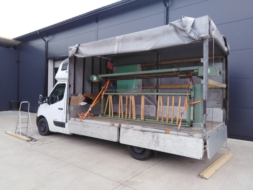 Biały bus z otwartą przestrzenią ładunkową, zabezpieczona pomarańczowymi pasami maszyna przemysłowa w środku, drabina oparta o bok pojazdu, betonowy plac.