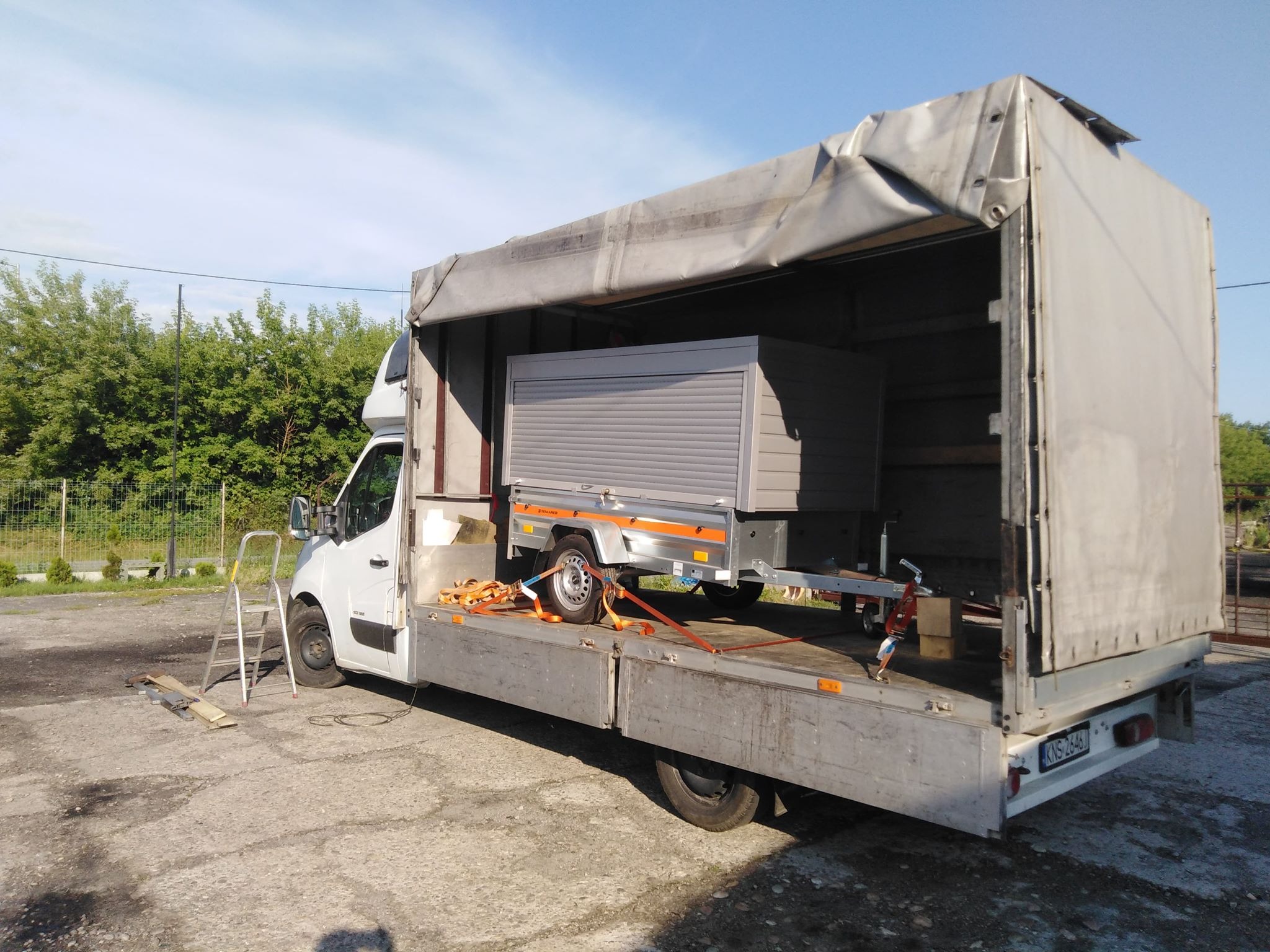 Przyczepa ze zwiniętą roletą zabezpieczona pasami na platformie samochodu dostawczego z otwartą plandeką, drabina oparta o bok pojazdu.