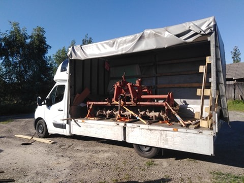 Biały bus z otwartą plandeką, przewożący czerwony sprzęt rolniczy zabezpieczony pasami transportowymi.