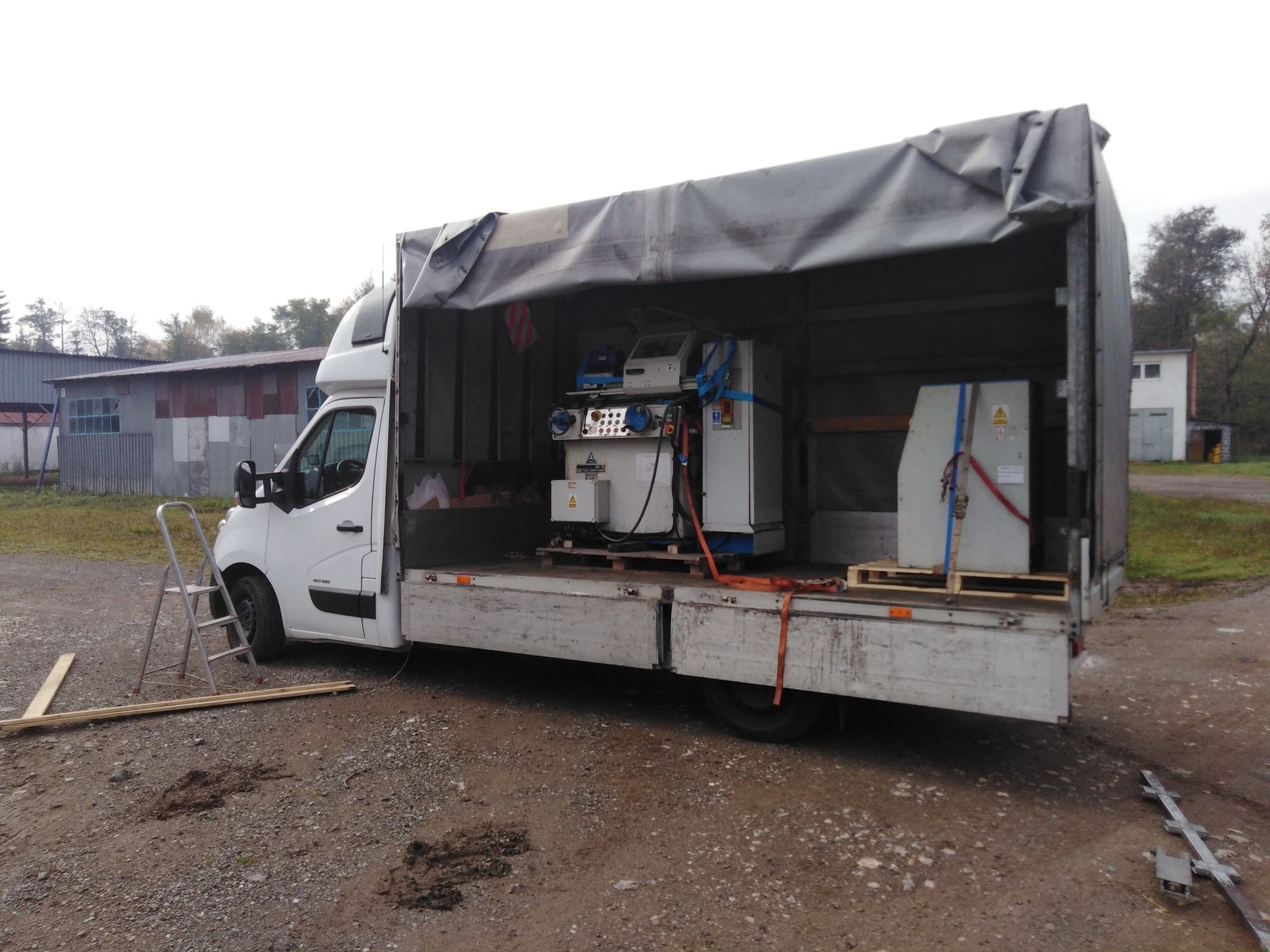 Biały bus z otwartą plandeką, wewnątrz zabezpieczone maszyny przemysłowe na paletach, drabina oparta o bok pojazdu, teren żwirowy.
