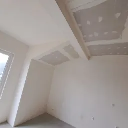 Wnętrze w trakcie remontu: surowy stan ścian i sufitu z widocznymi płytami kartonowo-gipsowymi przed szpachlowaniem, widoczne puszki elektryczne na ścianie, surowa wylewka podłogowa