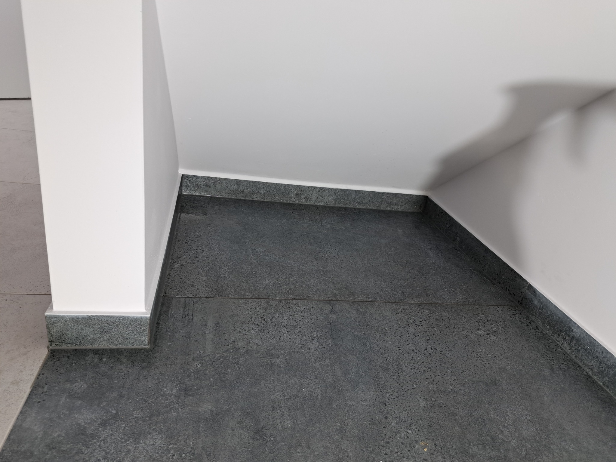 Szare, teksturowane płytki podłogowe z cokołem przy białej ścianie, widoczne łączenia płytek.