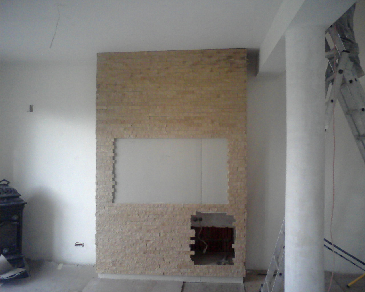 Ściana w trakcie wykończenia z dekoracyjną okładziną kamienną wokół miejsca na kominek i otworu na instalacje, widoczna drabina malarska.