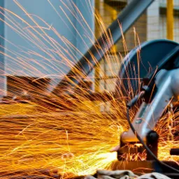 Pracownik w rękawicach tnący metal szlifierką kątową, z intensywnym snopem iskier i znakiem ewakuacyjnym w tle.