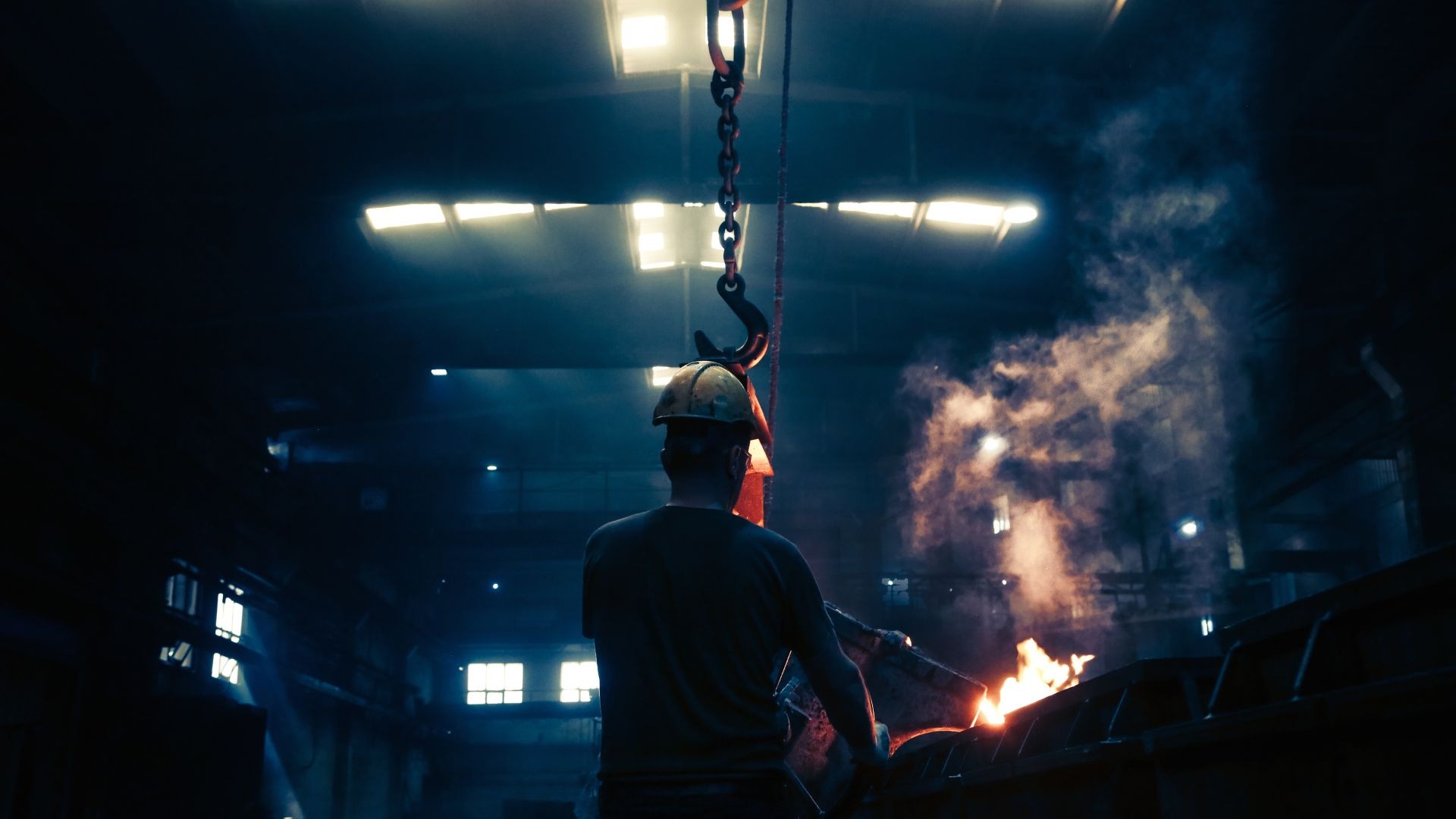 Pracownik w kasku obsługuje odlewniczy proces z użyciem suwnicy, widoczne płomienie i dym w słabo oświetlonej hali produkcyjnej.