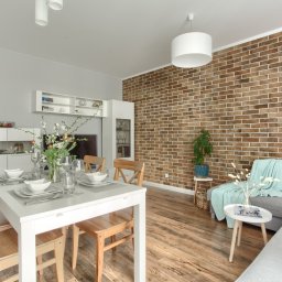 M2Photo Karolina Ibraheem - Jasne, przestronne wnętrze jadalni i salonu z ceglaną ścianą, drewnianą podłogą i nowoczesnymi meblami, w tym białym stołem z nakryciem, szarą sofą z turkusowym pledem i roślinami w doniczkach.