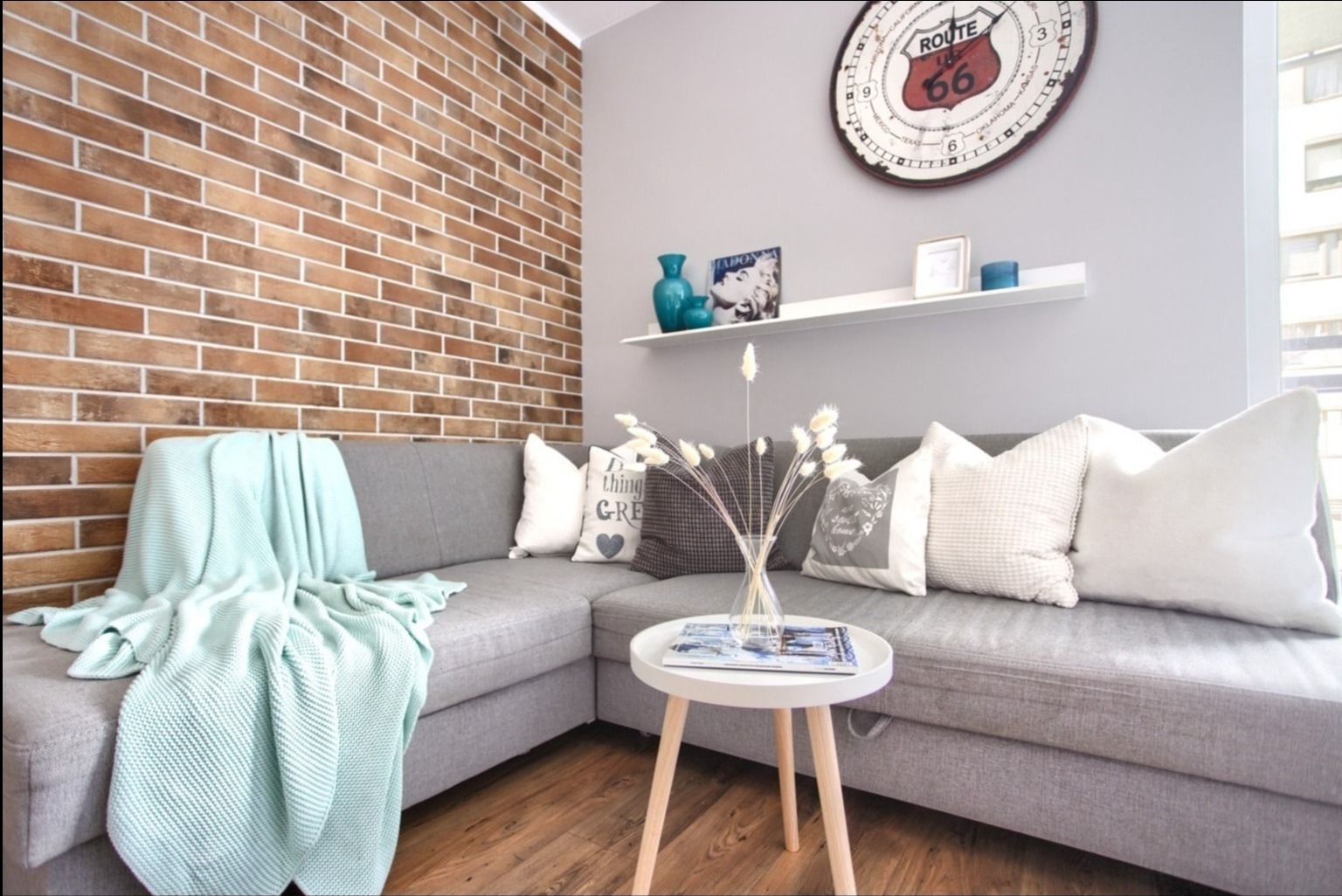 Salon z szarą narożną sofą, ceglaną ścianą, dekoracjami na półce i zegarem Route 66