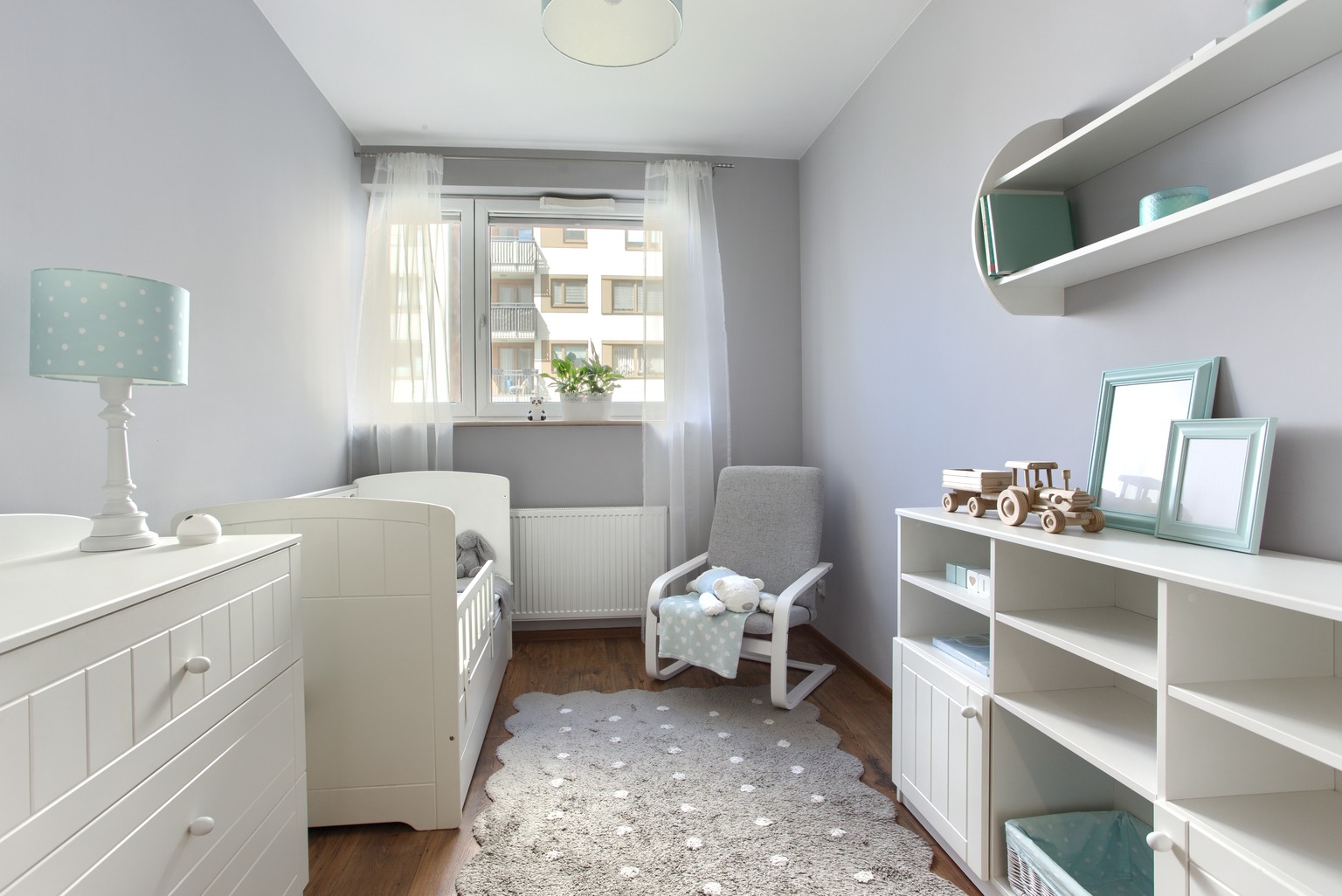Jasnoszary pokój dziecięcy z białymi meblami, łóżeczkiem, komodą, regałem i dywanem w kształcie owcy.