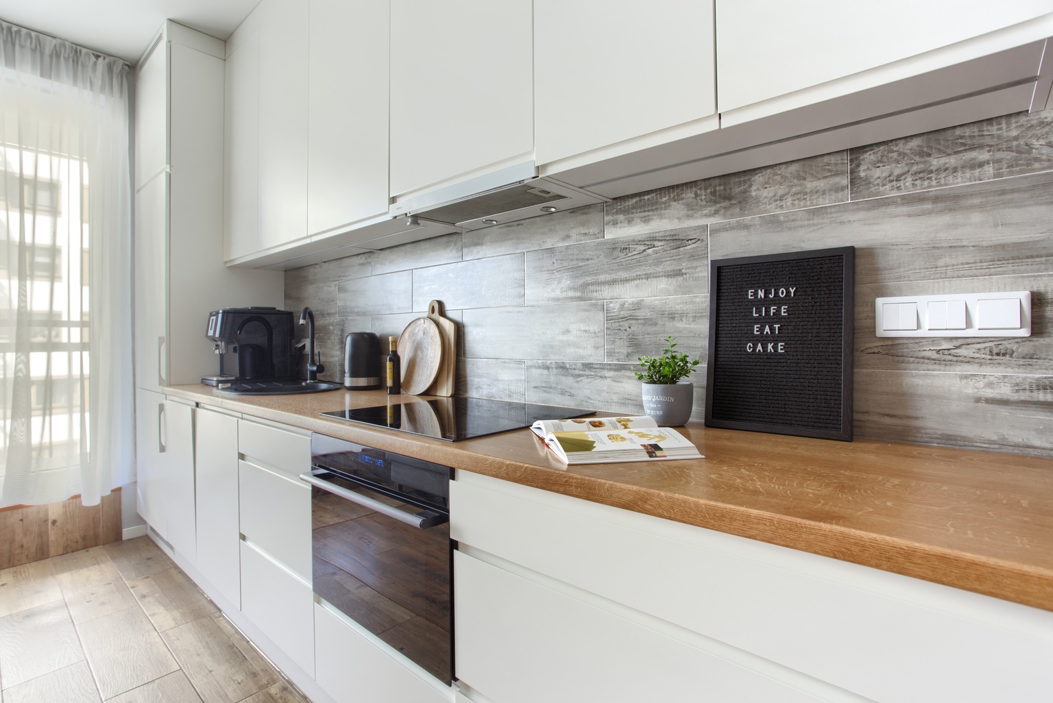 Jasna kuchnia z drewnianym blatem, białymi szafkami i szarymi płytkami imitującymi drewno. Na blacie ekspres do kawy, czajnik, deski do krojenia, płyta indukcyjna, piekarnik, książka kucharska...