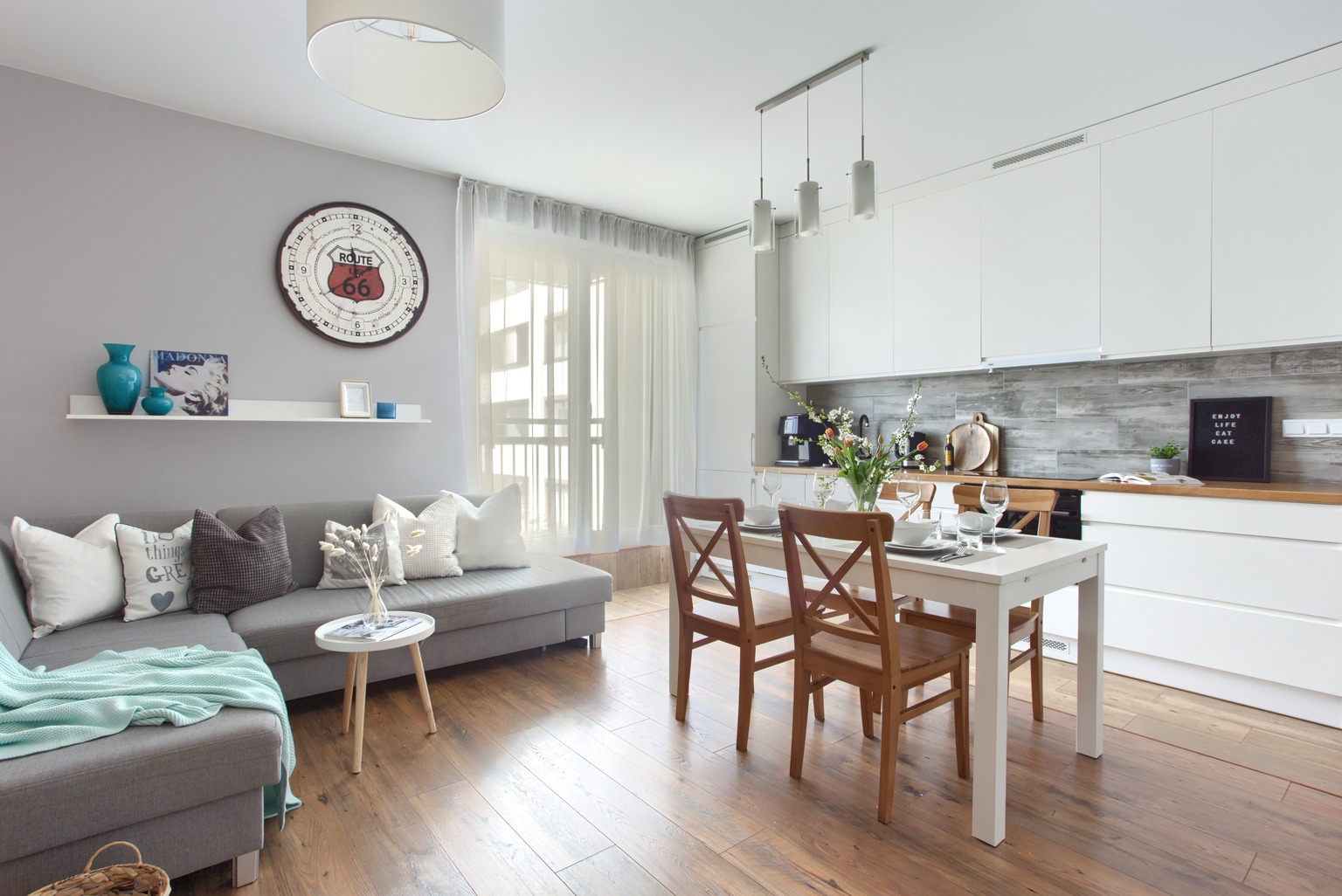 Jasne wnętrze mieszkania z szarą sofą, drewnianym stołem jadalnym i białą kuchnią, akcentowane dekoracjami w odcieniach błękitu i brązu.
