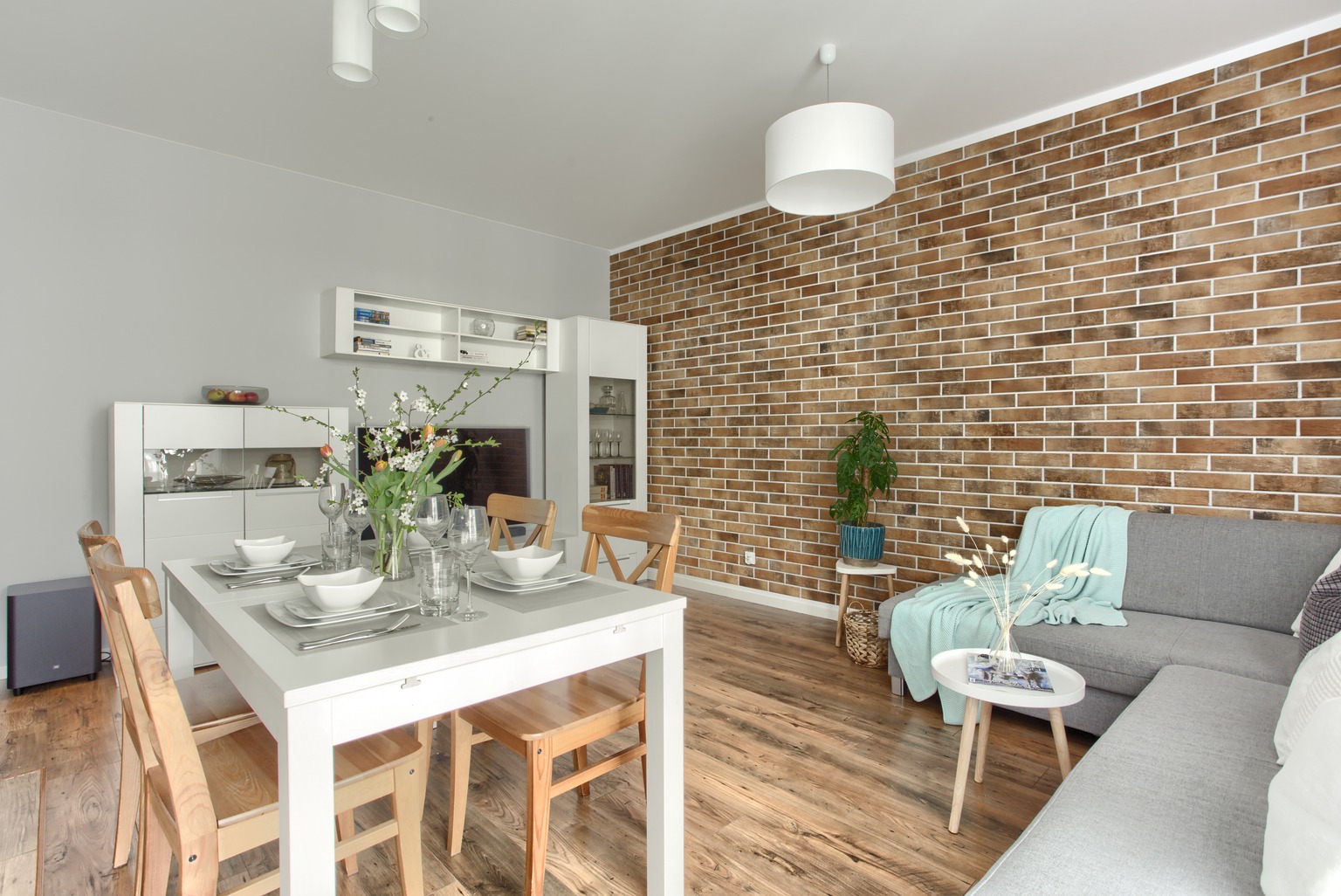 Jasne, przestronne wnętrze jadalni i salonu z ceglaną ścianą, drewnianą podłogą i nowoczesnymi meblami, w tym białym stołem z nakryciem, szarą sofą z turkusowym pledem i roślinami w doniczkach.