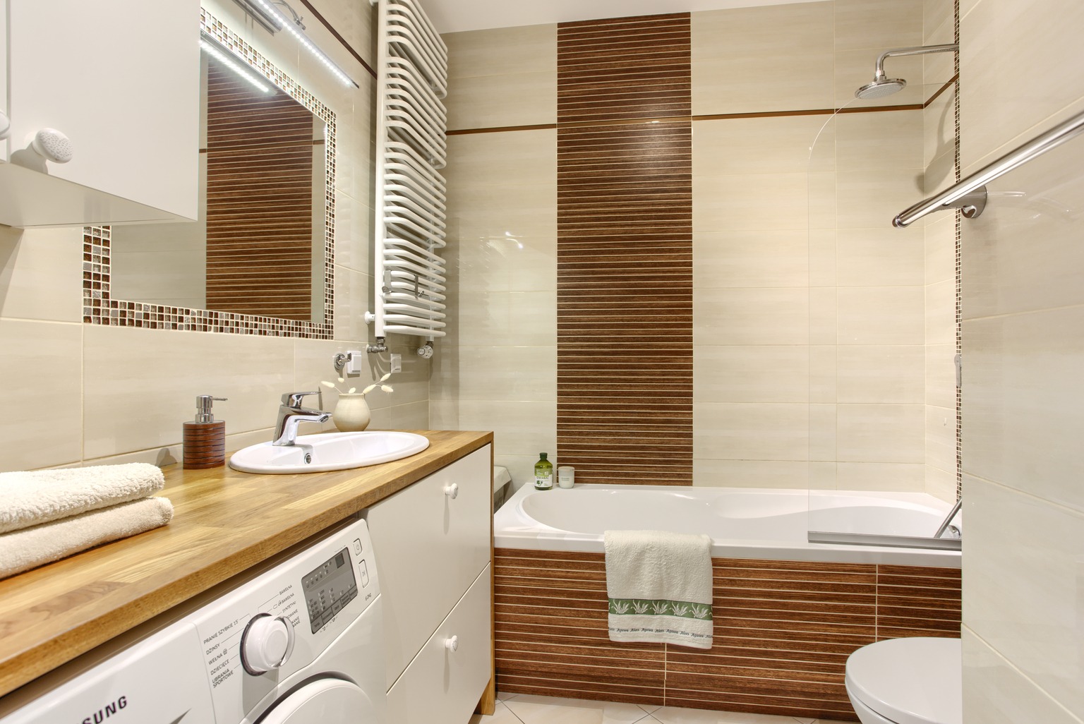 Jasna łazienka z drewnianym blatem, pralką, wanną z brązowymi akcentami i mozaikowym lustrem.
