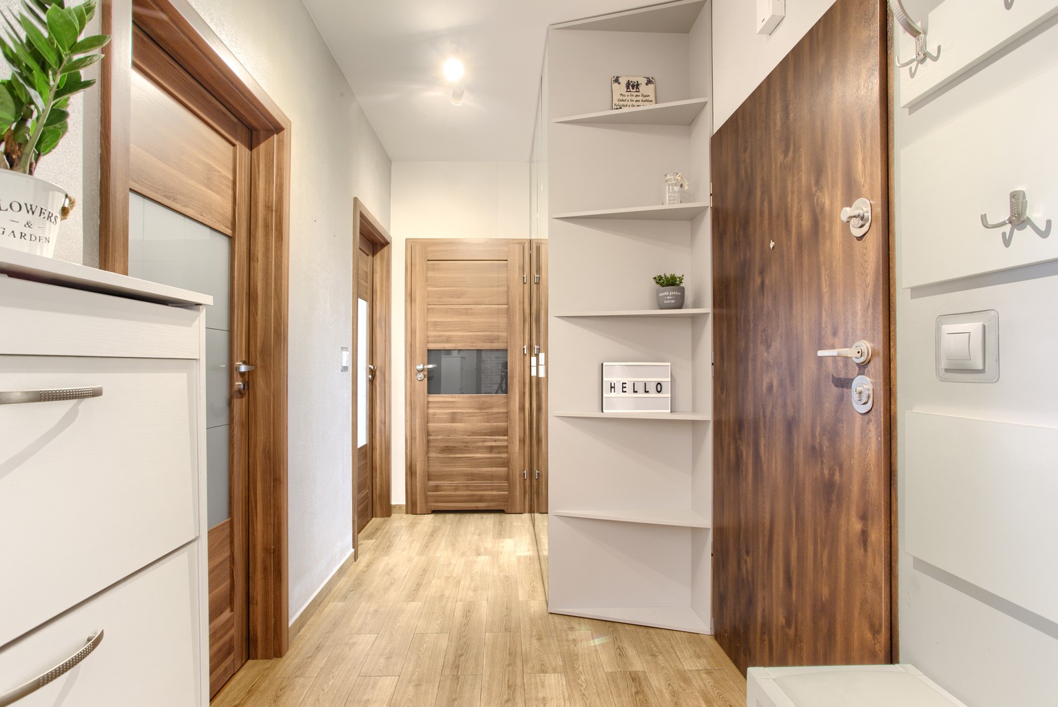 Jasny korytarz z drewnianymi drzwiami, białymi meblami i regałem z dekoracjami, w tym podświetlanym napisem 'Hello'.
