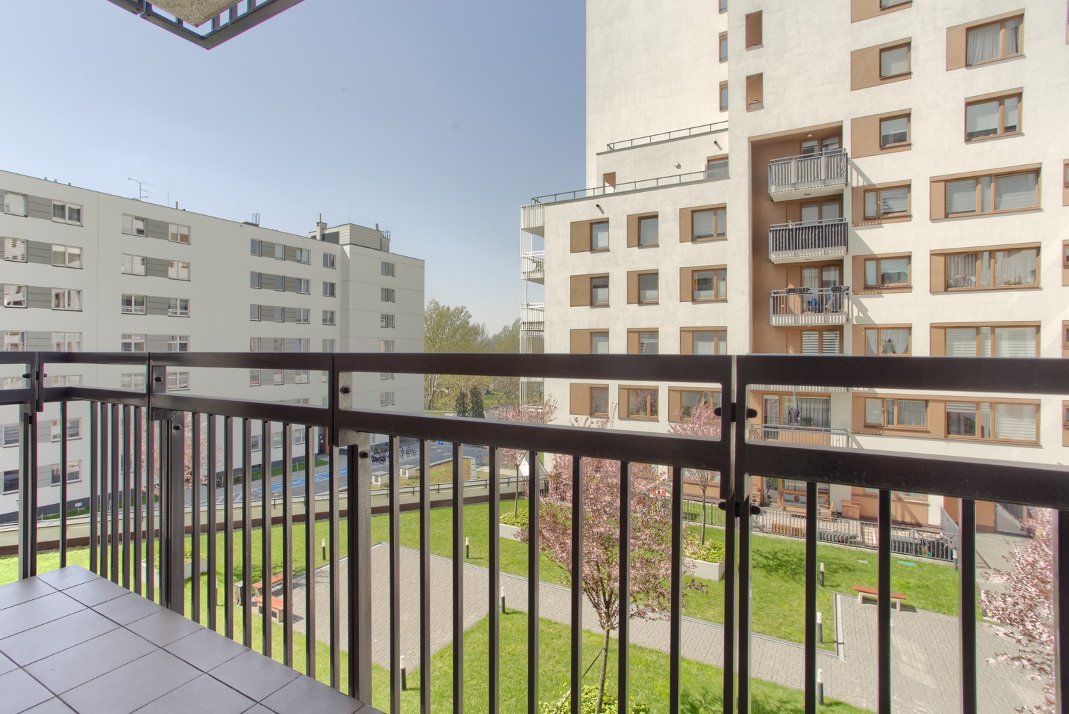 Widok z balkonu na osiedle mieszkaniowe z zadbanym trawnikiem i kwitnącymi drzewami, barierka balkonu w kolorze grafitowym.