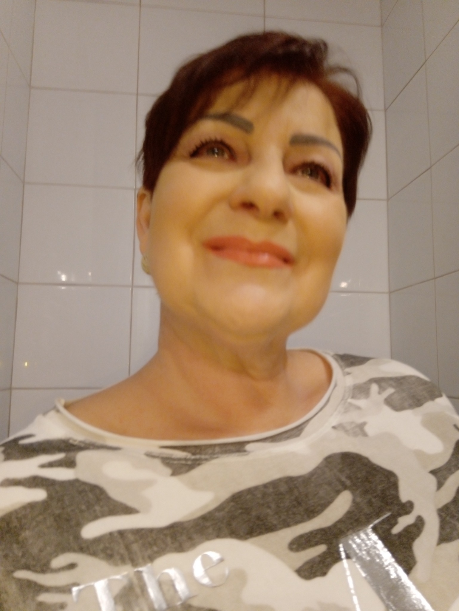 Portret kobiety w koszulce moro na tle kafelków, uśmiechnięta, z krótkimi brązowymi włosami i makijażem.