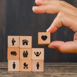 Drewniane kostki z ikonami: rodzina, dom, serce. Ręka układa kostkę z sercem na stosie, symbolizującym bezpieczeństwo i ochronę życia, rodziny, domu.