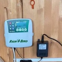 Sterownik nawadniania Rain Bird ESP-RZXe zamontowany na drewnianej ścianie, z zasilaczem i pomarańczowym kluczem wiszącym powyżej.