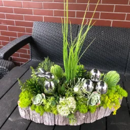 Dekoracja wielkanocna na drewnianym podkładzie z mchu, sukulentów, trawy i srebrnych jajek oraz zajączka na tle ściany z czerwonej cegły i czarnego plecionego fotela.