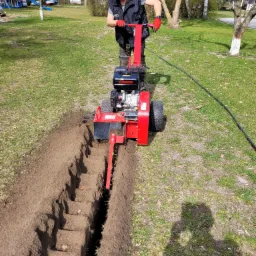 Mężczyzna w słuchawkach ochronnych obsługuje czerwoną koparkę łańcuchową, wykopując wąski rów w trawniku; widoczny cień operatora i fragment kabla zasilającego.