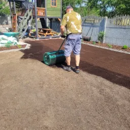 Mężczyzna wyrównuje glebę walcem ogrodowym na przygotowanym terenie pod trawnik, w tle drewniany plac zabaw dla dzieci i betonowe ogrodzenie.