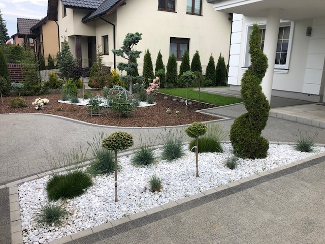 Starannie zaaranżowany ogród przed domem z geometrycznie przyciętymi krzewami, białymi kamieniami i korą, widoczne różne gatunki roślin, w tym formowane tuje spiralne i kule, oraz trawnik w tle.