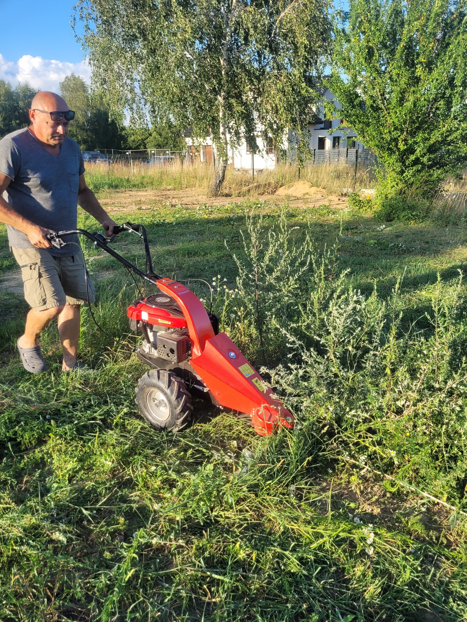 Mężczyzna w okularach słonecznych, ubrany w szarą koszulkę i beżowe szorty, prowadzi czerwoną kosiarkę listwową po zarośniętej trawie na tle domu i drzew.