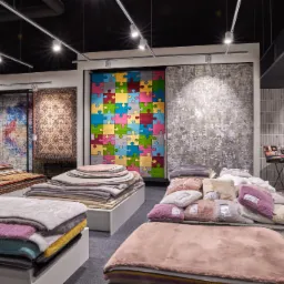 Wystawa dywanów w sklepie: różnorodne wzory, od tradycyjnych po nowoczesne, w tym dywan-puzzle, ułożone na białych podestach pod oświetleniem szynowym, z logo 'arte' na tle ściany z płytek.